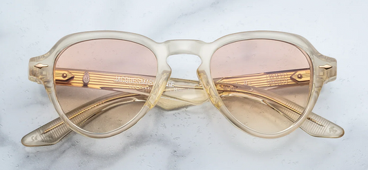 Front view of Jacques Marie Mage Hatfield sunglasses from the Photochromic Collection in Halo. The frames exhibit a compact, structured, retro-inspired aviator silhouette with a distinctively tailored lens shape. The heavy 10mm block acetate is a translucent Polished Brut champagne-beige, housing responsive photochromic brown gradient lenses. Signature Light Gold arrowhead front pins ornament the upper outer corners of the frame face. Available at WINK Optometrists.