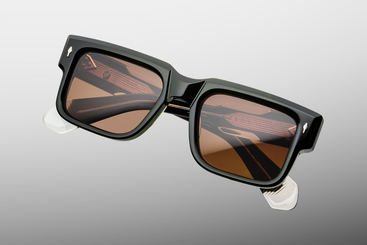 Close-up detail of the front corner of Jacques Marie Mage Hemmings Borneo sunglasses. Captures the polished, high-gloss finish of the earthy green and brown marbled acetate alongside a flush-mounted, gleaming gold arrowhead pin. WINK OPTOMETRISTS