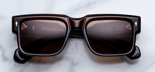 Front view of Jacques Marie Mage Hemmings Café sunglasses. Features a bold, boxy 10mm cured cellulose acetate frame in deep espresso brown, fitted with warm hazel-tinted lenses and signature gold arrowhead front pins.