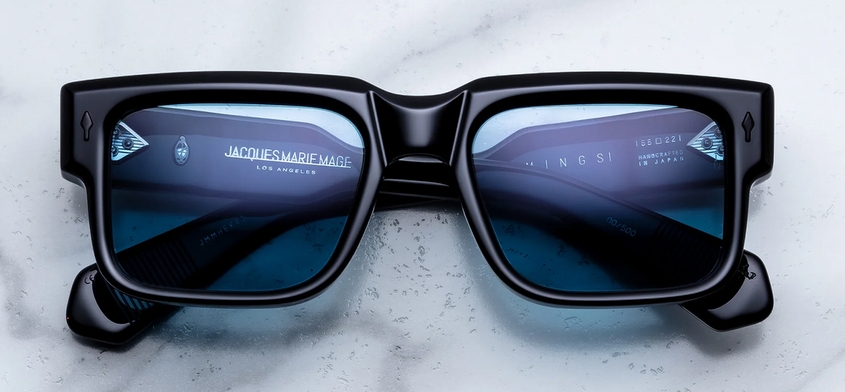A close-up studio photograph of Jacques Marie Mage Hemmings sunglasses in the limited-edition Mystique colourway. The frames feature a blocky, 1960s-inspired rectangular silhouette with a dual-laminated structure, displaying a matte black outer front paired with a polished, water-clear transparent inner layer. The deep blue, sapphire-toned lenses provide a stark, cool contrast to the dark frame. Signature arrowhead pins in a matching matte black finish are embedded at the outer temples.