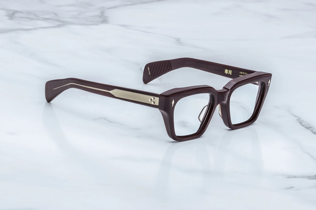 Three-quarter studio shot of the Jacques Marie Mage Ichikawa Wine RX glasses. The perspective highlights the heavy 10mm profile depth of the rich red acetate front and its cleanly sculpted, architectural edges. Clear demo lenses are bordered by the thick-gauge trapeze rims, seamlessly blending into custom dual-laminated matching wine and clear acetate temples. Available at WINK Optometrists
