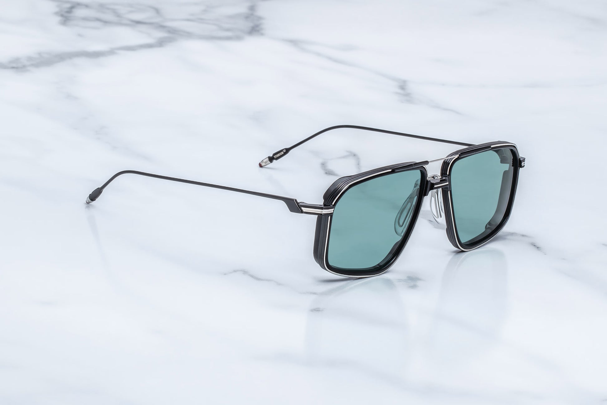 Three-quarter studio shot of the JMM Jagger Paon sunglasses, showcasing the geometric black double bridge, structural brow bar, and the vibrant peacock-blue lens tint.