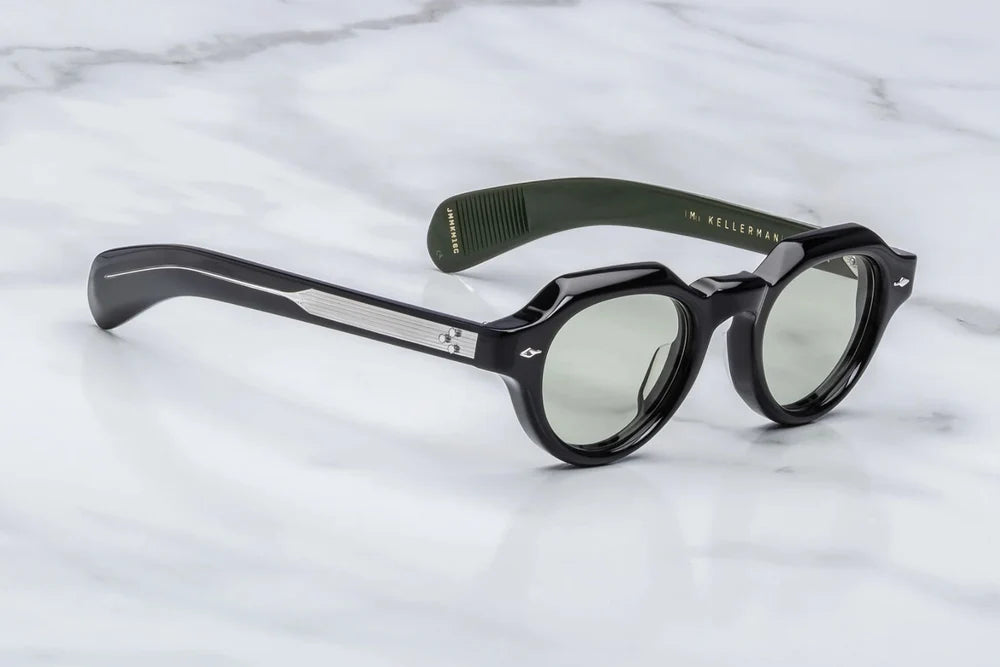 Three-quarter studio shot of the Jacques Marie Mage Kellerman Cobra glasses. The perspective highlights the heavy 10mm profile depth of the glossy black frame face and its skillfully sculpted, faceted bevels. The clear demo lenses highlight the structural thickness of the rims as they seamlessly blend into custom double-laminated black and green acetate temples. Available at WINK Optometrists.
