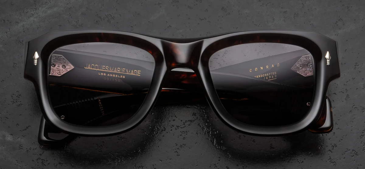 Front view of Jacques Marie Mage Conrad Beau sunglasses. Features a bold, 10mm thick Havana tortoiseshell acetate frame with an architectural, beveled square silhouette. Sterling silver signature arrowhead pins adorn the top outer corners, housing solid green-grey Stratus mineral glass lenses