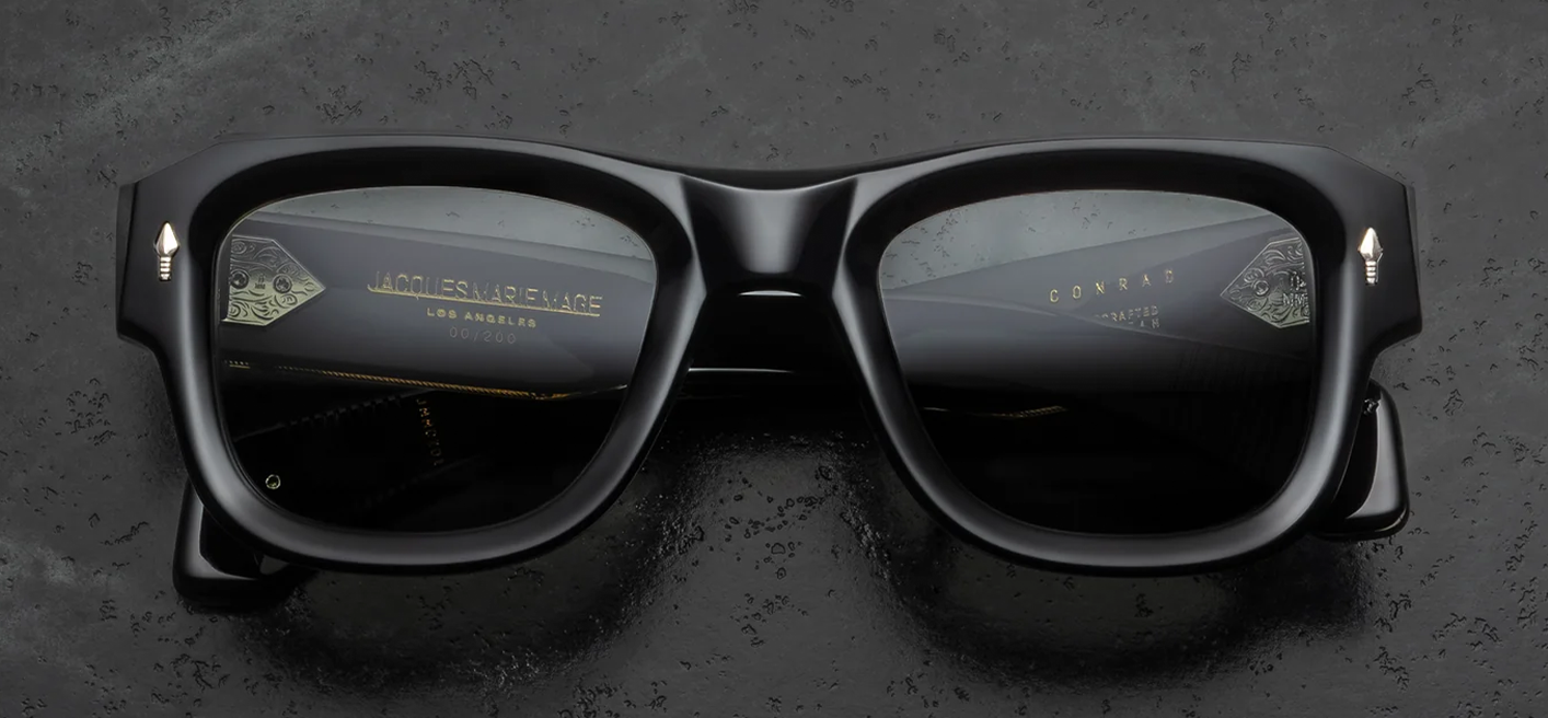  Front view of Jacques Marie Mage Conrad Noir sunglasses. Features a thick, 10mm black acetate frame with a boldly beveled, squared silhouette. Signature sterling silver Last Frontier arrowhead pins sit at the top outer corners, housing neutral grey 4-base mineral glass lenses.
