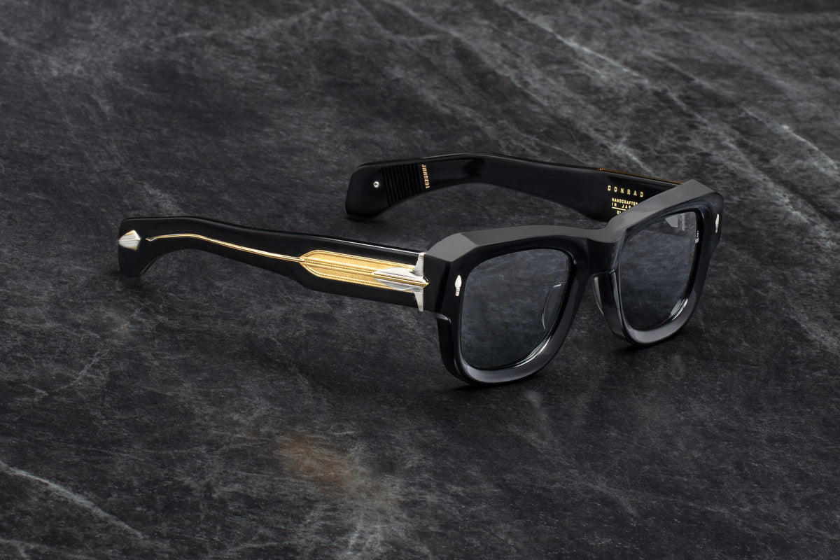  Three-quarter studio shot of Jacques Marie Mage Conrad Noir sunglasses. The angle highlights the deep structural beveling on the heavy black acetate frame and showcases the dramatic transition from the solid black front frame to the transparent, gold-core temple arms.