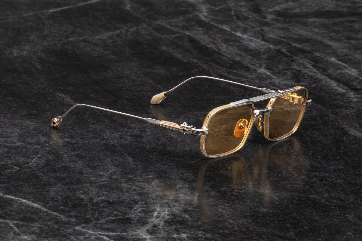 Three-quarter studio shot of Jacques Marie Mage Deaver Electrum sunglasses, highlighting the contrast between the silver palladium frame, dark gold details, and yellow lenses. The perspective reveals the hairline engraving details along the top bar and the gemstone-inlaid arrowhead hinge ornament