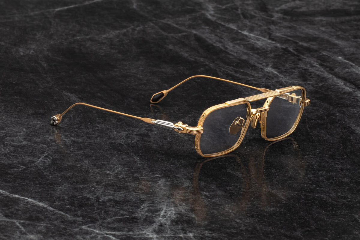 Three-quarter studio shot of Jacques Marie Mage Deaver Maxi sunglasses, showcasing the dark gold frames and grey Harbor lenses. The angle highlights the precision hairline engravings on the flat top bar and the contrast of the silver-and-onyx arrowhead hinge ornament