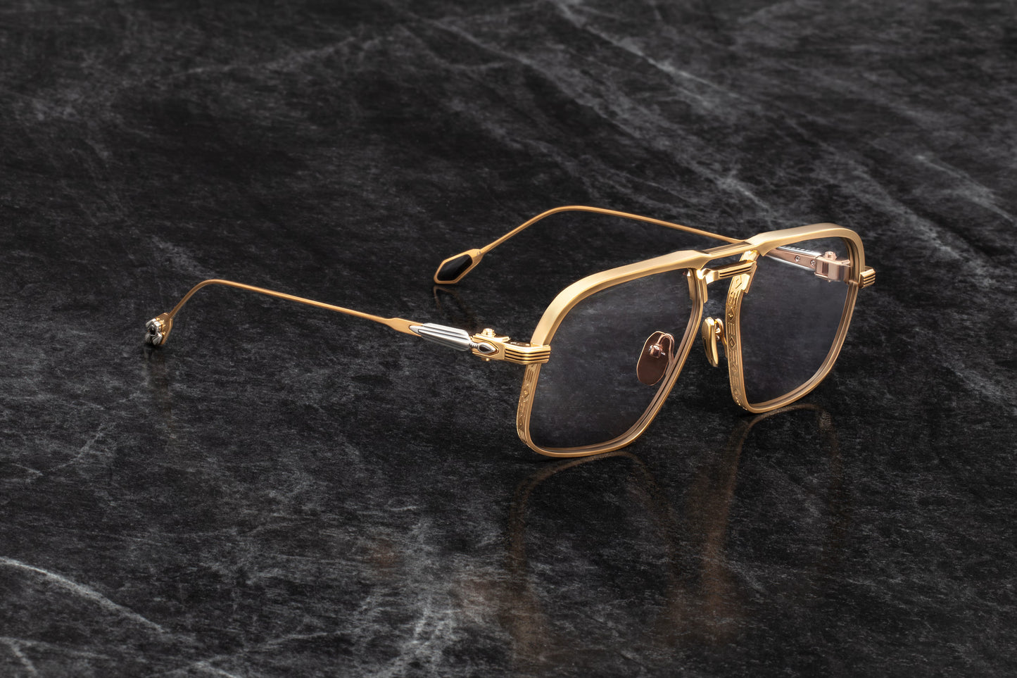 Three-quarter studio shot of Jacques Marie Mage Labarge Maxi sunglasses, showcasing the dark gold frames and solid grey lenses. The perspective highlights the hairline engravings on the thick top bar and the contrast of the sterling silver and black onyx arrowhead temples