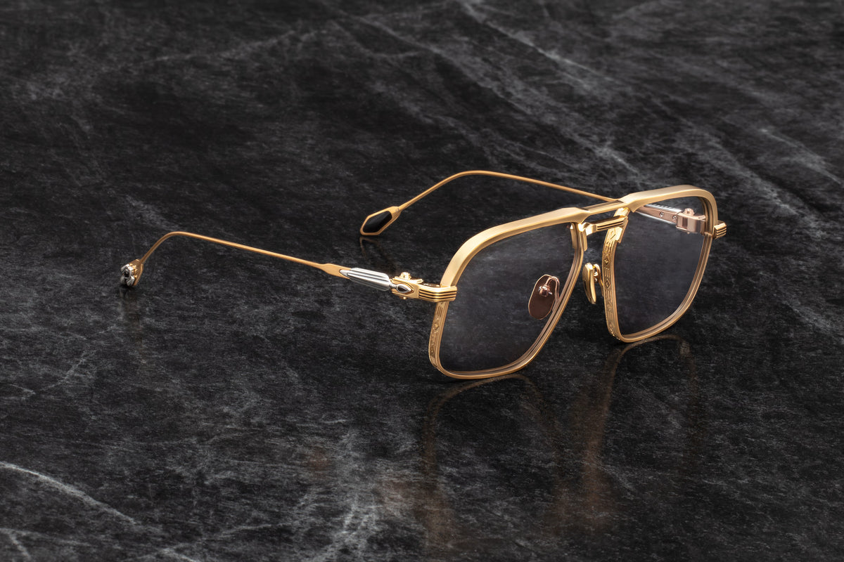 Three-quarter studio shot of Jacques Marie Mage Labarge Maxi sunglasses, showcasing the dark gold frames and solid grey lenses. The perspective highlights the hairline engravings on the thick top bar and the contrast of the sterling silver and black onyx arrowhead temples
