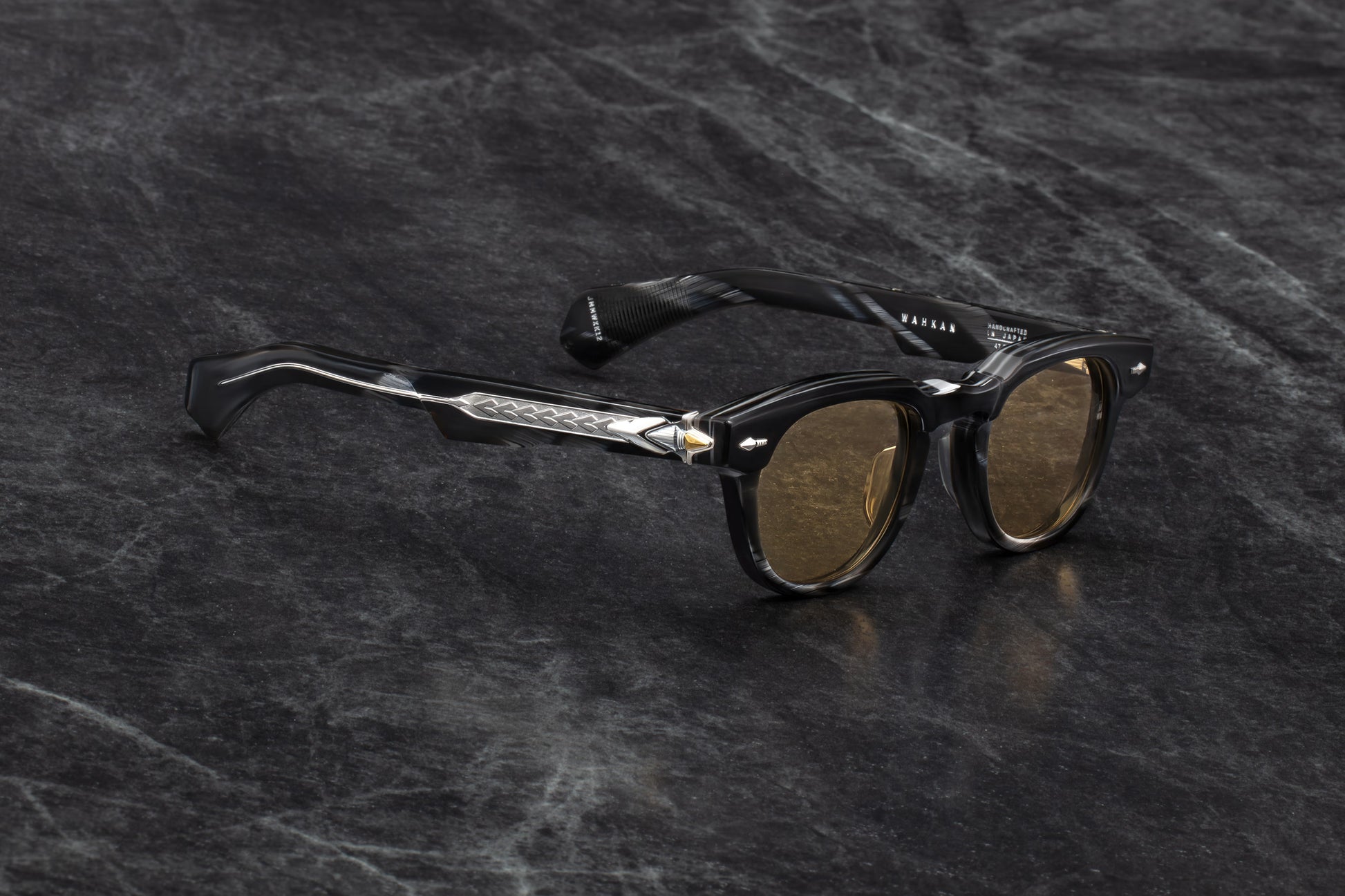 Three-quarter studio shot of Jacques Marie Mage Wahkan Ridgeway sunglasses. The perspective showcases the heavy beveling of the polished black horn frame and the contrast against the clear temple arms that reveal the metallic wirecore. The warm honey lenses catch the light, showing their subtle curvature