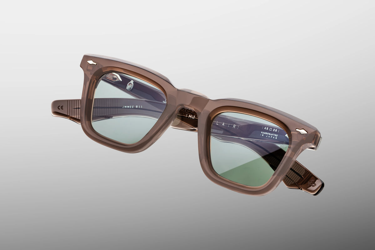 A macro close-up of the front corner of the JMM Leclair Dusty Jade frame. The shot details the premium polished finish of the translucent grey-taupe acetate. It highlights the signature sterling silver arrowhead front pin embedded perfectly flush into the frame, with a glimpse of the rich green lens catching the light.