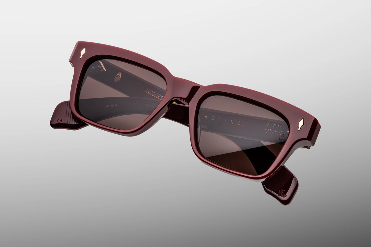 Extreme close-up of the front corner of Jacques Marie Mage Molino Bordeaux Empire sunglasses. Highlights the rich, translucent quality of the polished burgundy acetate, smooth beveled contours, and a flush-mounted sterling silver arrowhead pin.