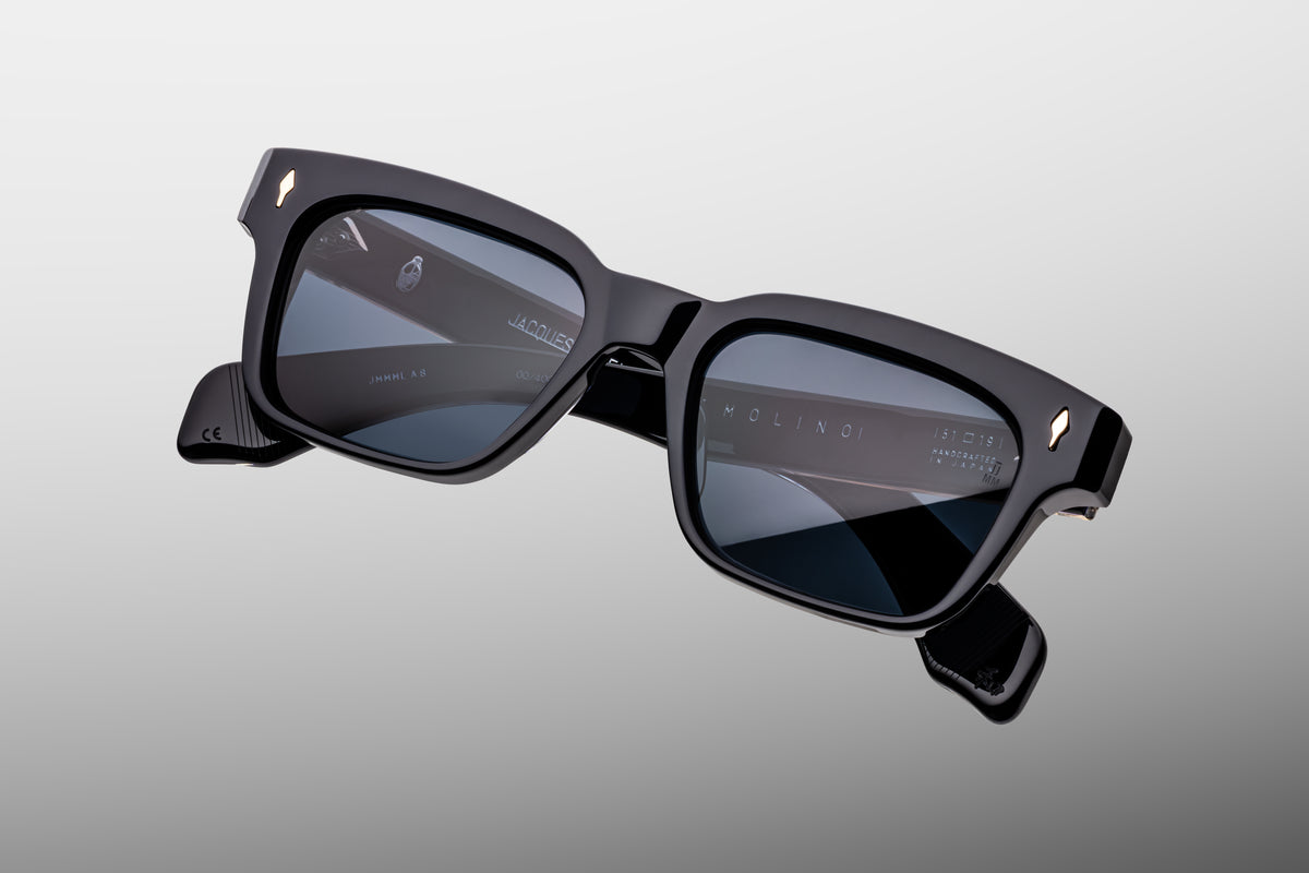 Extreme close-up of the front corner of Jacques Marie Mage Molino Lacquer sunglasses. Captures the rich, high-gloss finish of the dark lacquer acetate, sharp bevel details, and a flush-mounted, polished light-gold arrowhead pin.