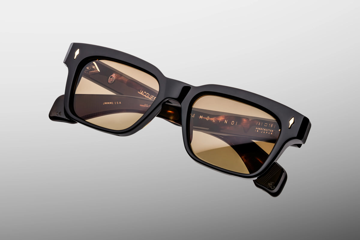 Extreme close-up of the front corner of Jacques Marie Mage Molino Noir 15 sunglasses. Captures the high-gloss shine of the black block acetate frame, smooth beveled contours, and a flush-mounted sterling silver arrowhead pin.