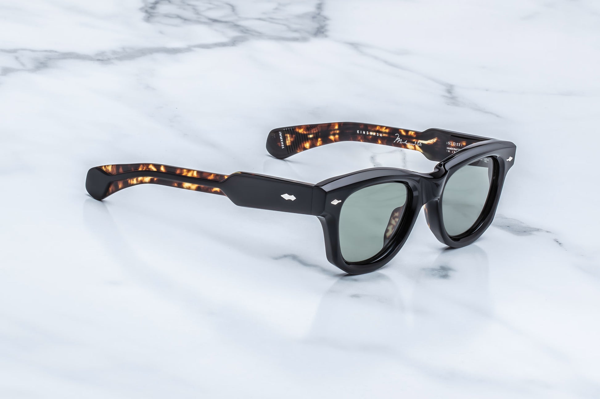 Three-quarter studio shot of the JMM Kinshasa in Noir, showcasing the thick 10mm block acetate frame, dark Sequoia green lenses, and the bold, sculptural front-to-temple transition