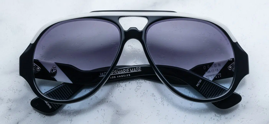 A close-up of the Jacques Marie Mage Orion sunglasses in Navy, featuring a bold, '80s-inspired aviator silhouette with a deep teardrop lens shape. The frame is crafted from 7mm cured navy blue cellulose acetate, complemented by white brow and temple details. The lenses are Moon Dance gradient grey CR39 with backside anti-reflective coating, offering 100% UV protection. 