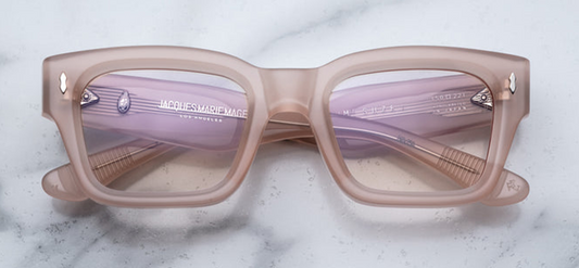 Straight-on view of Jacques Marie Mage Suze in Pink Quartz: a bold, medium‑size rectangular eyeglass frame made from 10 mm cured light-pink cellulose acetate. The full‑rim silhouette has gently softened square corners and a slightly raised saddle bridge. Superlight pink-tinted CR‑39 lenses with 4-base curvature are flush set. Each upper front corner features a sterling‑silver arrowhead pin, adding a refined metallic accent against the soft pastel frame.