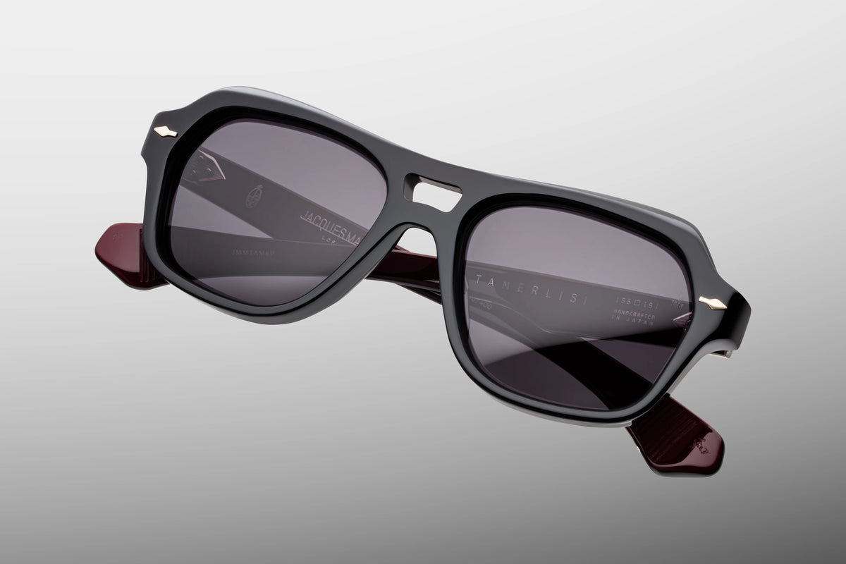 Extreme close-up of the front chassis on Jacques Marie Mage Tamerlis Bloodstone sunglasses. Captures the high-gloss shine of the black block acetate, the complex multi-angled beveling along the arched brow, and a flush-mounted sterling silver arrowhead pin.