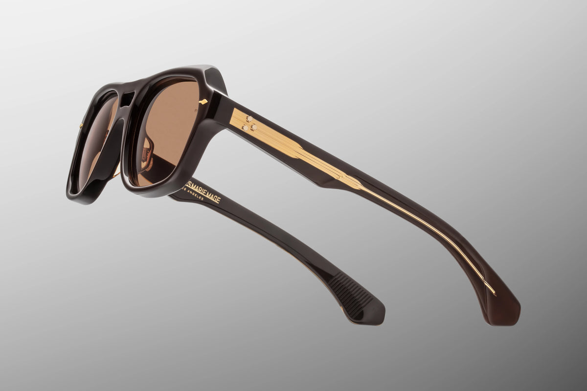 Macro shot of the temple arm on Jacques Marie Mage Tamerlis Walnut sunglasses. The crystal-clear beige outer laminate layer acts as a window to the exposed dark gold Lever wire core engraved with intricate hairline filigree.