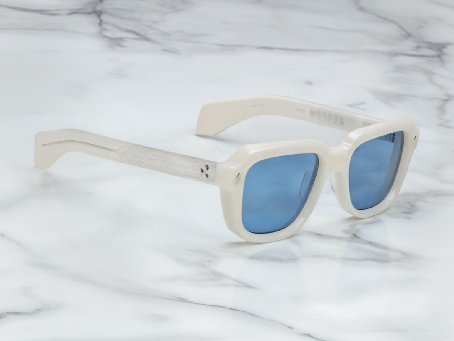 Side view of Jacques Marie Mage Taos in Mykonos sunglasses, bold, rectangular 10 mm white cured acetate frames evoking Mykonos’ architecture. They feature exposed silver wire‑core temples threaded with tribal-engraved detailing, vintage-style 4-base blue gradient CR39 lenses with backside anti‑reflective coating, and full UV protection. Inspired by Dennis Hopper’s eyewear in Easy Rider, this is a limited edition of 150 pieces at WINK Optometrists
