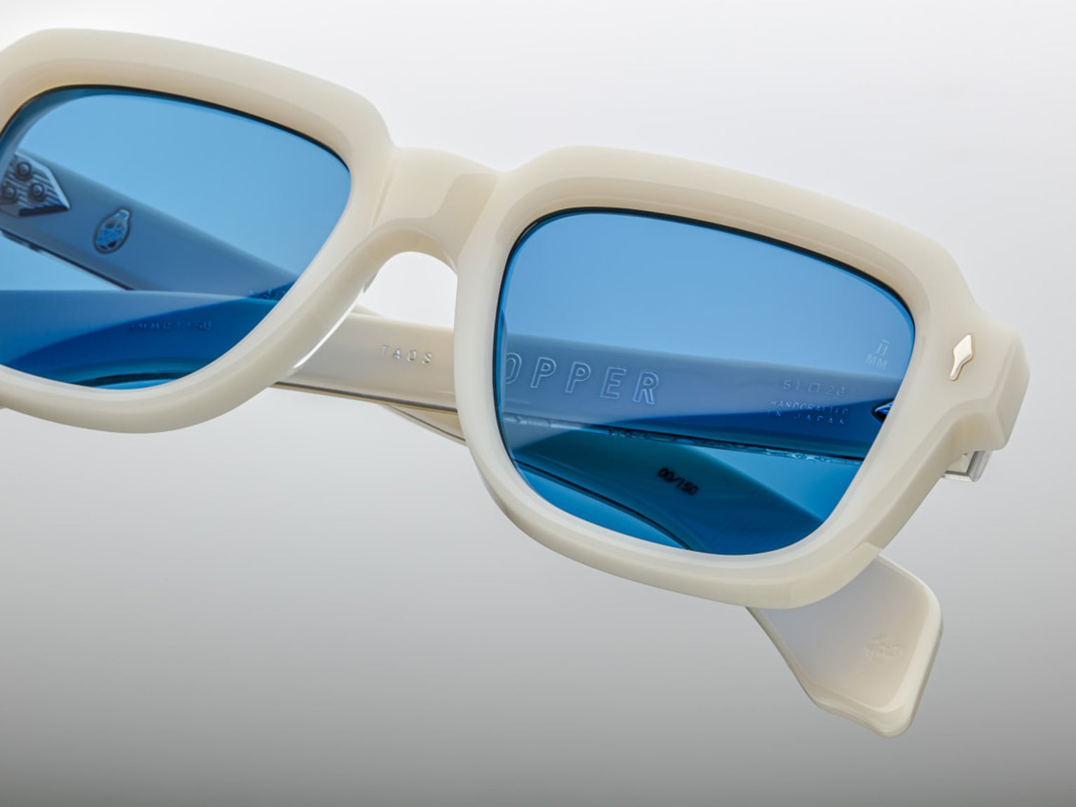 Close details of Jacques Marie Mage Taos in Mykonos sunglasses, bold, rectangular 10 mm white cured acetate frames evoking Mykonos’ architecture. They feature exposed silver wire‑core temples threaded with tribal-engraved detailing, vintage-style 4-base blue gradient CR39 lenses with backside anti‑reflective coating, and full UV protection. Inspired by Dennis Hopper’s eyewear in Easy Rider, this is a limited edition of 150 pieces at WINK Optometrists