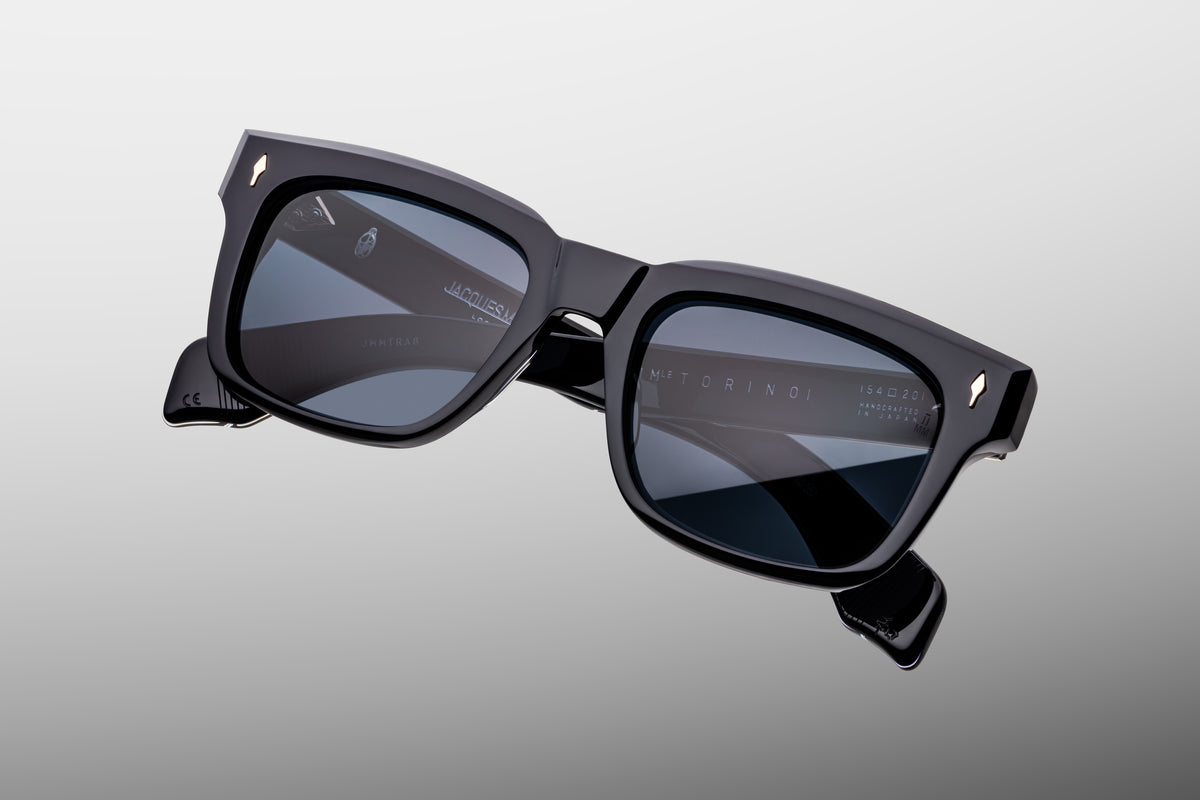 Extreme close-up of the front corner of Jacques Marie Mage Torino Lacquer sunglasses. Captures the high-gloss luster of the wet-ink dark blue acetate frame, smooth beveled contours, and a flush-mounted light-gold arrowhead pin.