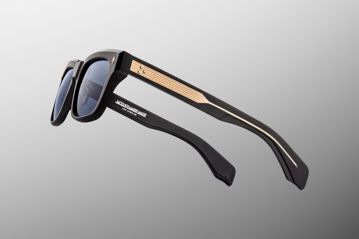 Macro shot of the temple arm on Jacques Marie Mage Torino Lacquer sunglasses, showcasing the subtle play of deep blue light against the frame's dark surface and the premium metallic hinge hardware.