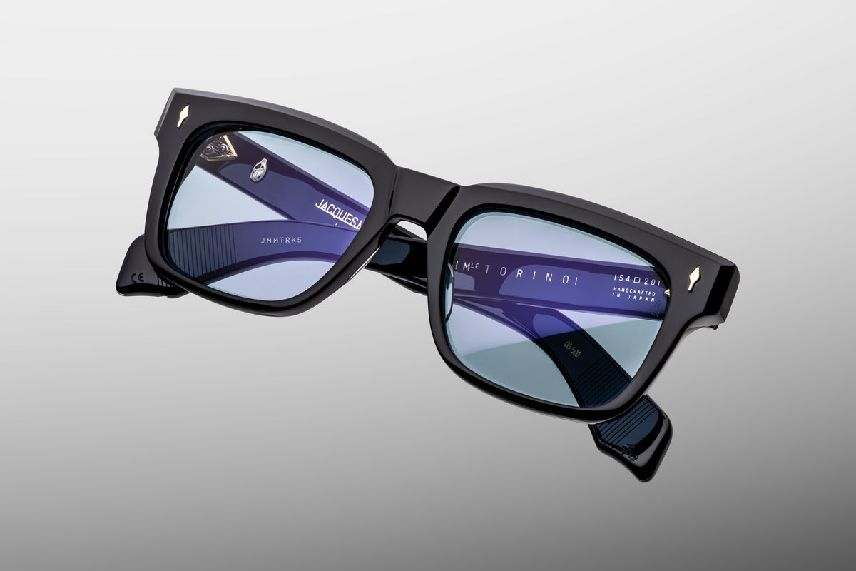 Extreme close-up of the front corner of Jacques Marie Mage Torino Obscure sunglasses. Captures the high-gloss shine of the black block acetate frame, smooth beveled contours, and a flush-mounted dark gold arrowhead pin.
