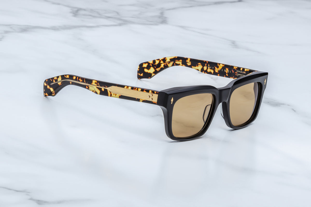Three-quarter view of the Jacques Marie Mage Torino Tavern sunglasses. The angle highlights the frame's structural volume, hand-polished angular beveling along the brow line, and thick temples that showcase a gold wire core through laminated clear interior acetate.