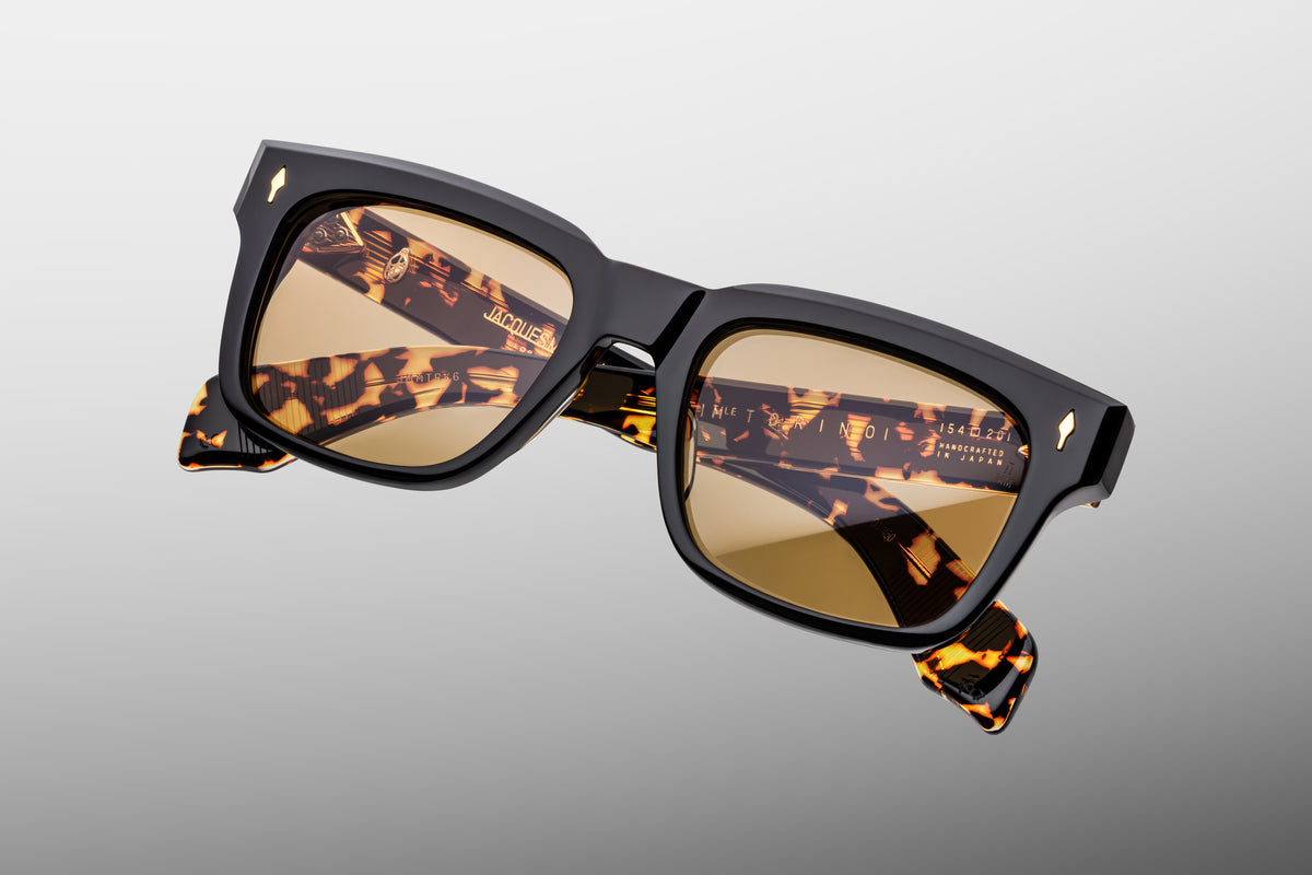 Extreme close-up of the front corner of Jacques Marie Mage Torino Tavern sunglasses. Captures the high-gloss shine of the warm marbled brown acetate frame, smooth beveled contours, and a flush-mounted dark gold arrowhead pin.