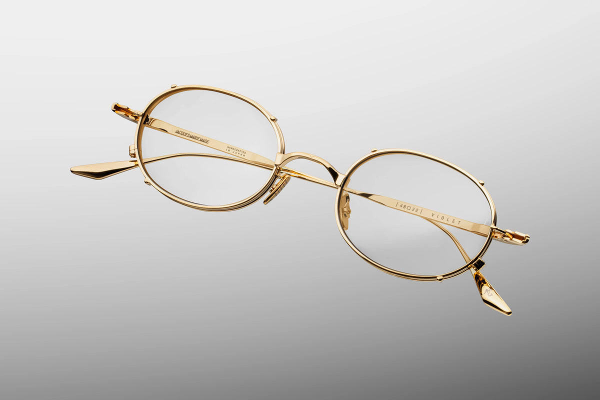 A macro close-up of the front outer corner of the JMM Violet RX frames. The image captures the theatrical craftsmanship of the dual-layered metal eye-rims and faux clip-on framework. It highlights the meticulous, fine lines of the premium titanium chassis enclosing the clear optical lens