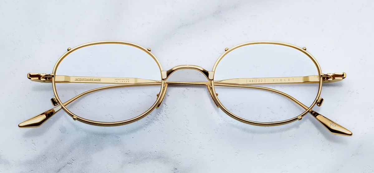 A front-facing product shot of Jacques Marie Mage Violet RX optical glasses. The frames feature a delicate, late-90s-inspired oval silhouette crafted from ultra-fine titanium. Clear prescription demo lenses sit inside a sculptural double eye-rim frame, accented by a distinctive architectural faux clip-on metal bridge
