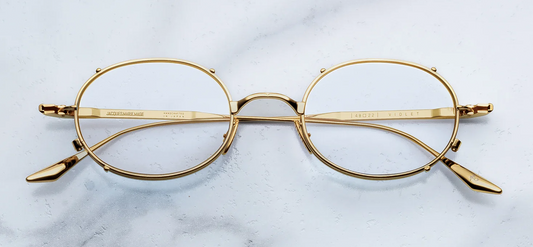 A front-facing product shot of Jacques Marie Mage Violet RX optical glasses. The frames feature a delicate, late-90s-inspired oval silhouette crafted from ultra-fine titanium. Clear prescription demo lenses sit inside a sculptural double eye-rim frame, accented by a distinctive architectural faux clip-on metal bridge