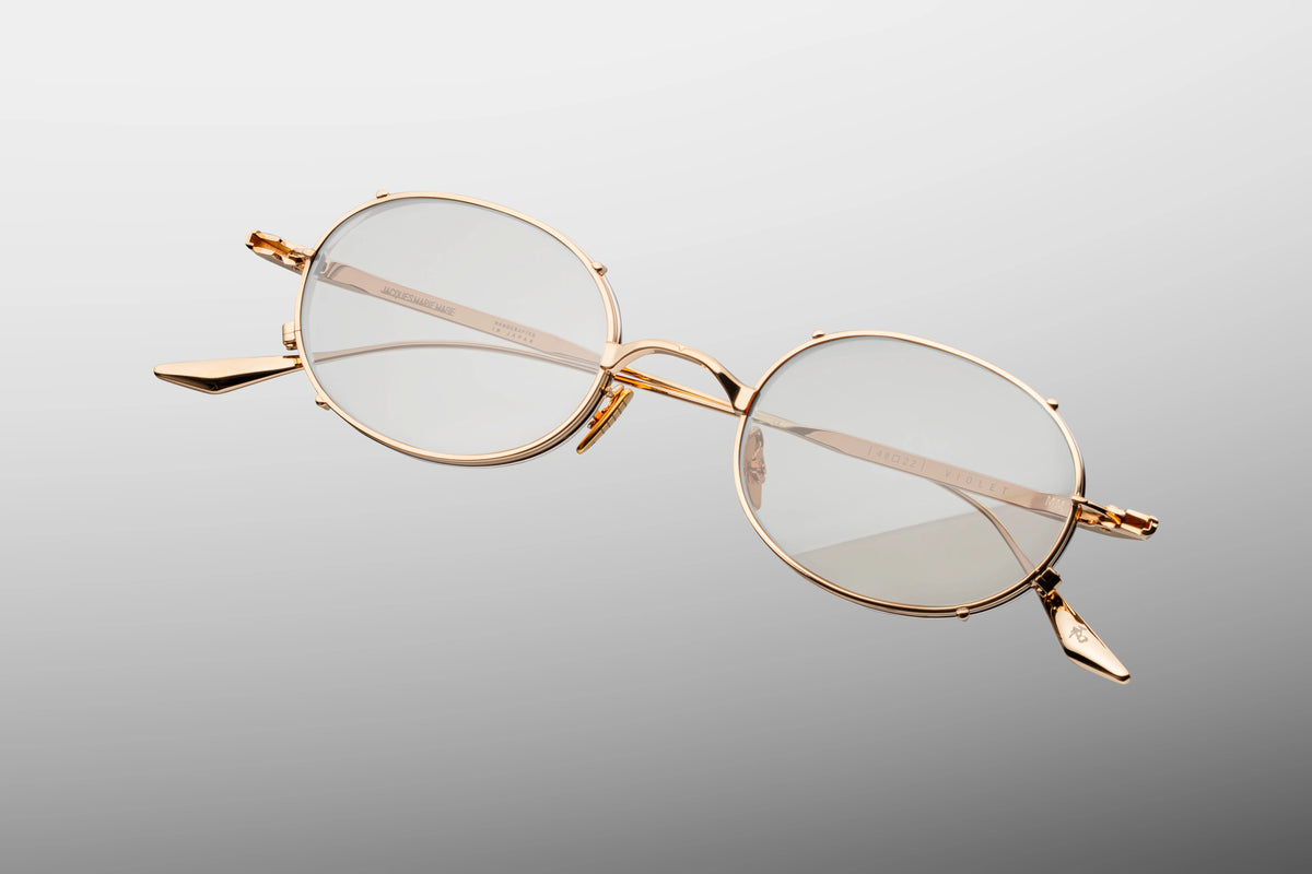 A macro close-up of the front outer corner of the JMM Violet RX Darjeeling frames. The image emphasizes the theatrical dual-layered gold and silver metalwork and the distinct faux clip-on bridge construction. Light reflects off the highly polished, meticulously sculpted titanium bezel enclosing the lens.