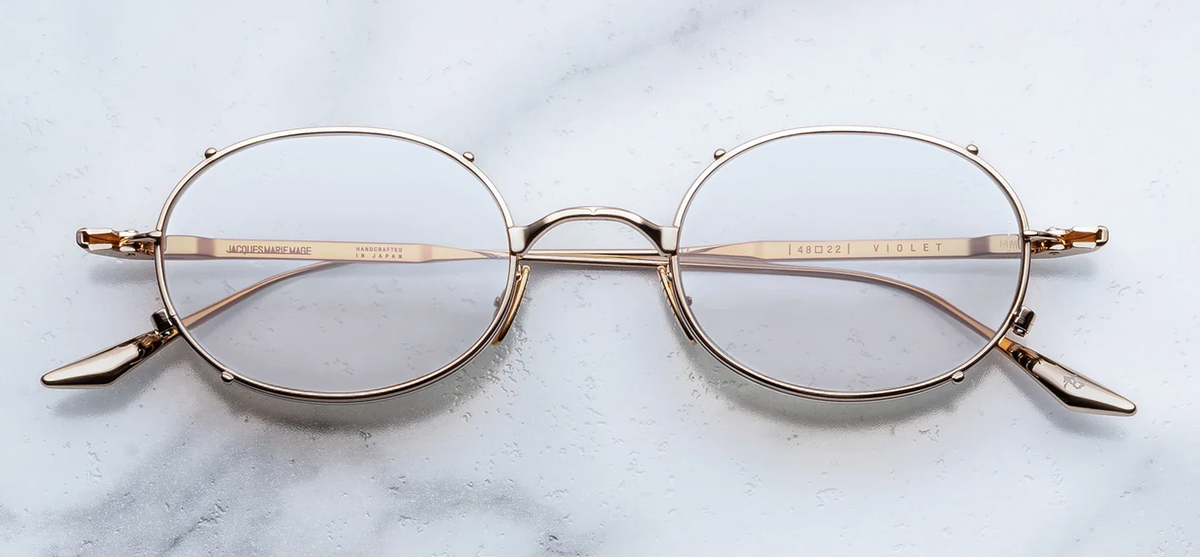 A front-facing product shot of Jacques Marie Mage Violet RX spectacles in the Darjeeling colourway. The minimalist, late-90s-inspired oval silhouette features a dual-tone premium beta-titanium structure with a polished silver-tone inner frame and a shimmering gold-tone outer layer. Clear lenses sit neatly inside the architectural double eye-rim structure