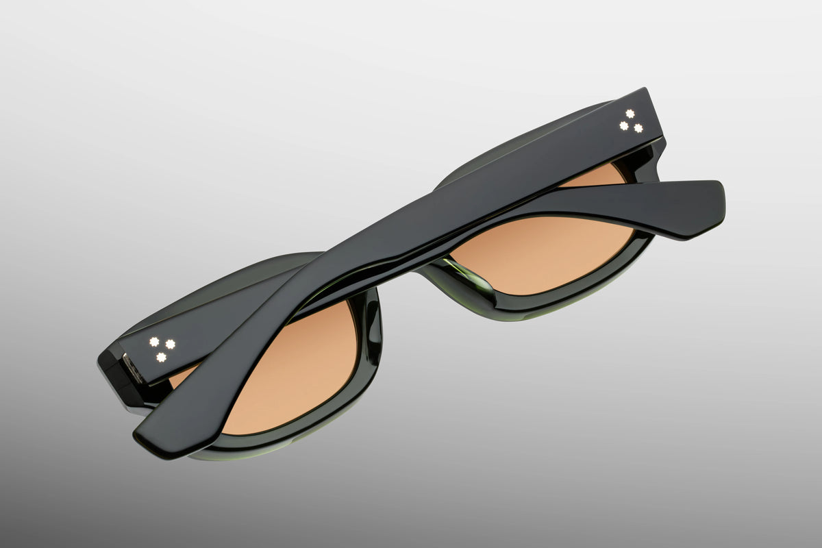 Detailed close-up view from behind the hinge of the JMM Whiskeyclone Bornéo sunglasses. The perspective displays a heavy-duty, custom 7-barrel tension-secured hinge with fine hairline details anchoring the temple arm to the green chassis. Foil-stamped branding text and a silver spur-shaped rivet are cleanly visible along the interior temple surface.
