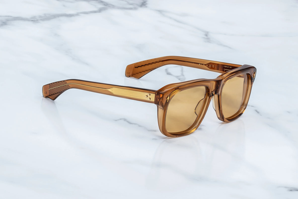 Three-quarter studio shot of the Jacques Marie Mage Yves Solaris photochromic sunglasses. The perspective emphasizes the heavy 10mm profile depth of the polished whiskey-colored frame face and its architectural, hand-polished contours. The adaptive photochromic orange lenses catch the light, flowing seamlessly into double-laminated temples with translucent amber interiors. Available at WINK Optometrists.