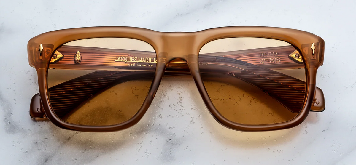 Front view of Jacques Marie Mage Yves sunglasses from the Photochromic Collection in Solaris. The frames present a classic 1970s rectangular silhouette with refined soft angles and notable volumes. The 10mm thick cured acetate block is a translucent Polished Whiskey Crystal amber-gold, housing responsive photochromic orange gradient lenses. Signature 18K Dark Gold arrowhead front pins ornament the upper outer corners. Available at WINK Optometrists.