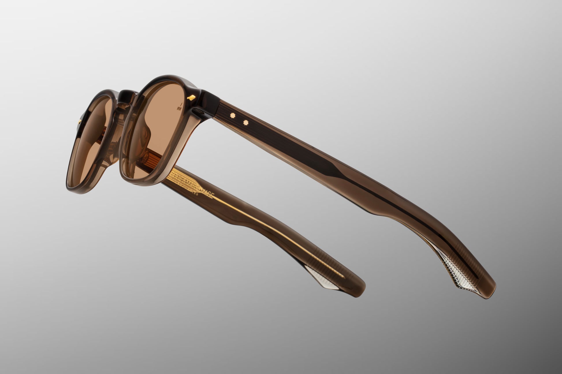 Side view of the JMM Zephirin London sunglasses temple arm, showcasing the glossy translucent brown outer temple, flush gold spur-shaped rivets, and the custom inner metal wirecore running through the structure.