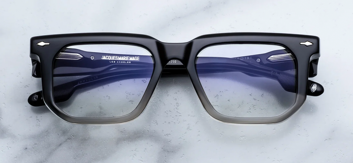 Front view of Jacques Marie Mage Yoshi RX eyeglasses in Black Fade. The frames present a low-profile geometric silhouette with a straight brow line. The upper half of the thick acetate frame face is a high-gloss jet black that seamlessly gradients into a crystal-clear translucent acetate along the lower rim. Signature sterling silver arrowhead pins accent the upper outer corners. Fitted with clear demo lenses. Available at WINK Optometrists.

