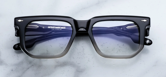 Front view of Jacques Marie Mage Yoshi RX eyeglasses in Black Fade. The frames present a low-profile geometric silhouette with a straight brow line. The upper half of the thick acetate frame face is a high-gloss jet black that seamlessly gradients into a crystal-clear translucent acetate along the lower rim. Signature sterling silver arrowhead pins accent the upper outer corners. Fitted with clear demo lenses. Available at WINK Optometrists.
